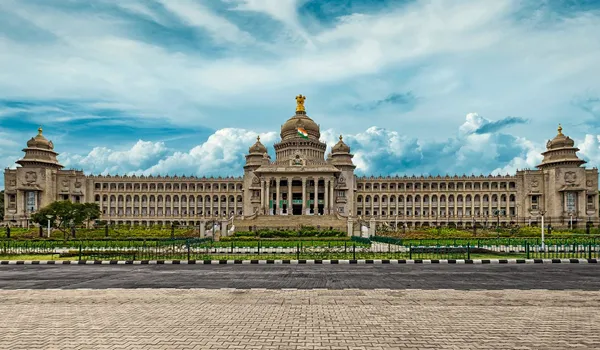 A panoramic cityscape view of Bangalore, showcasing a blend of modern high-rise corporate tech parks and a lush green tree canopy under a clear sky.